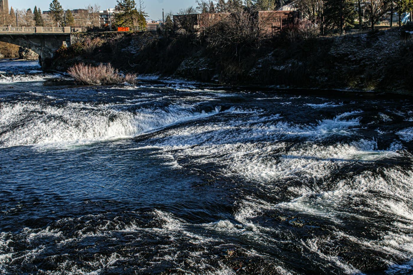Rapids on the Spokane River with a bridge and trees along the bank